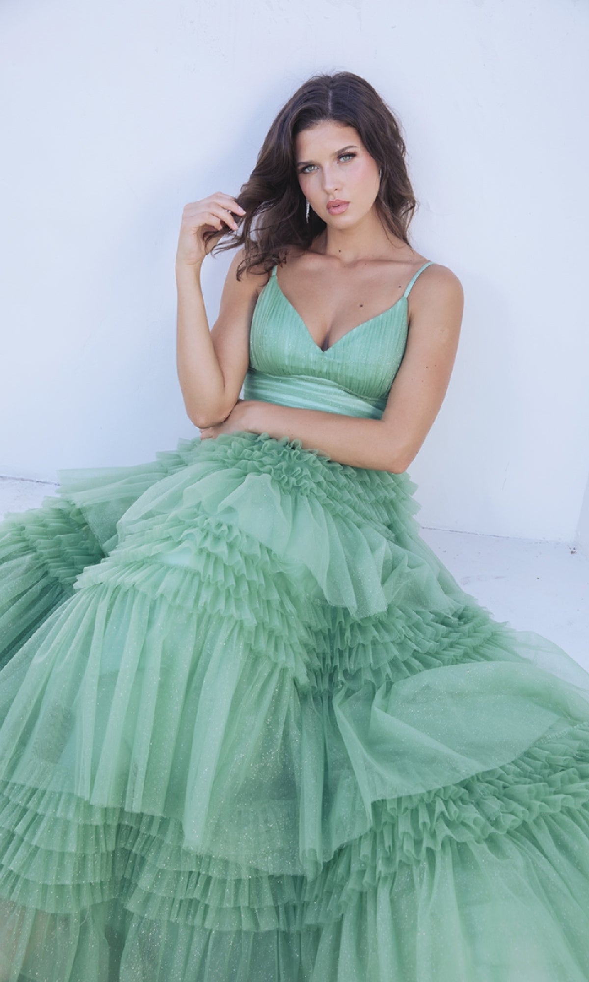 A woman in the Lace-Up Long Glitter-Tulle Prom Dress: Eureka 9888, a sleeveless green ruffled gown, sits against a white background, gazing at the camera with a neutral expression.